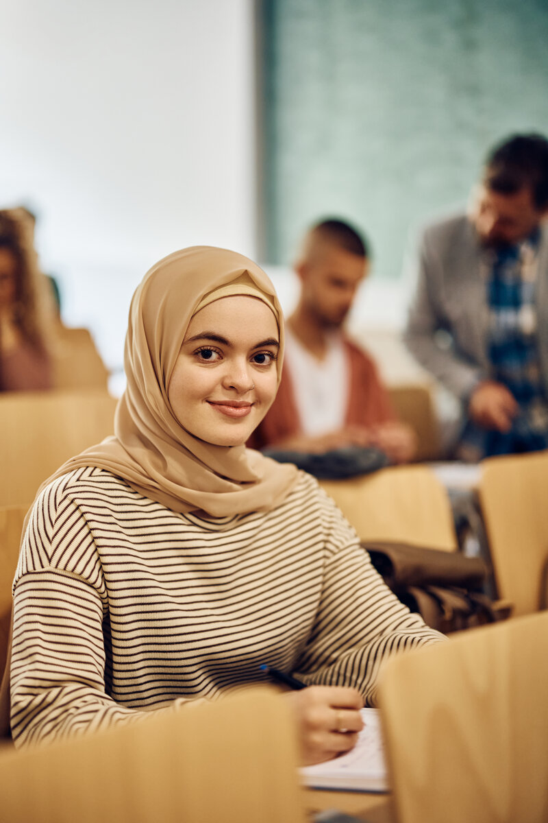 Young smiling Middle Eastern woman during lecture at college classroom looking at camera.